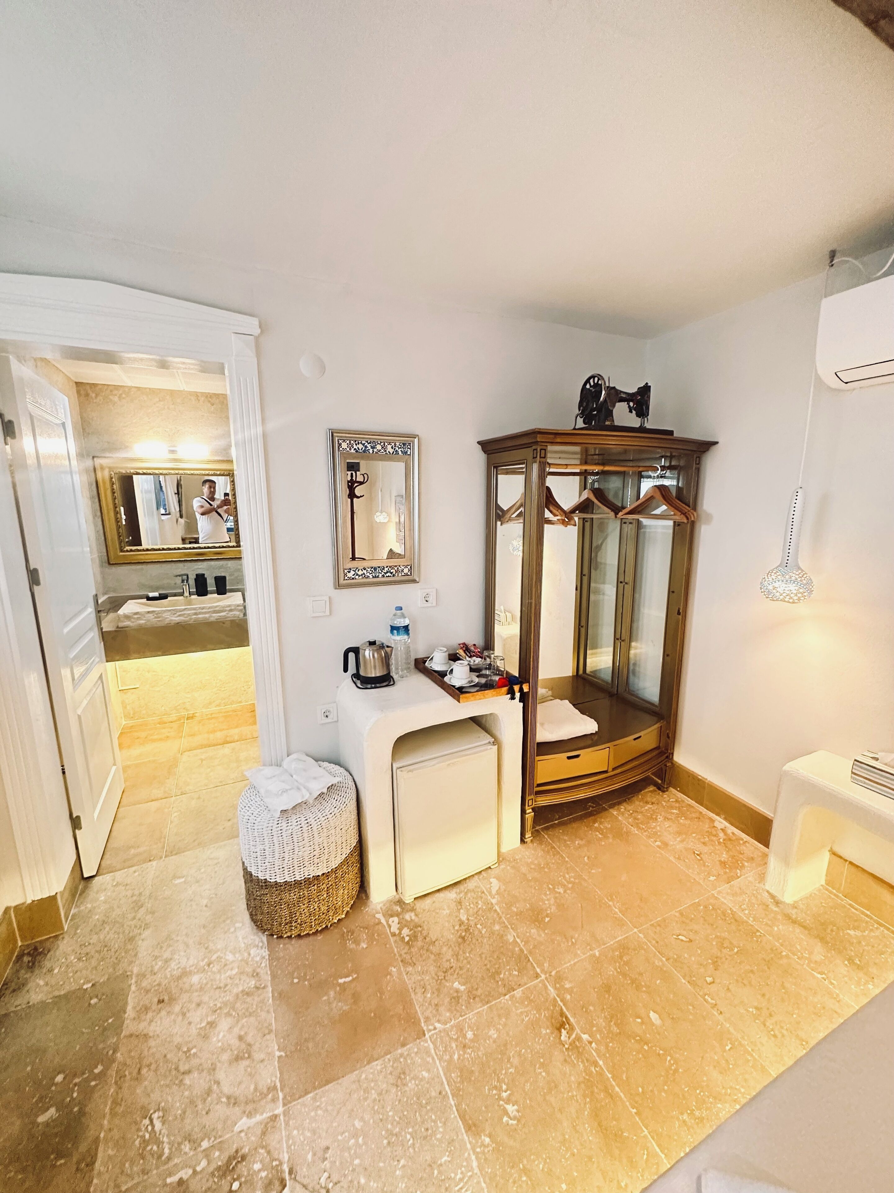 A compact hotel room with a small kitchenette, a vanity with a mirror, a wooden wardrobe, and a wicker stool.