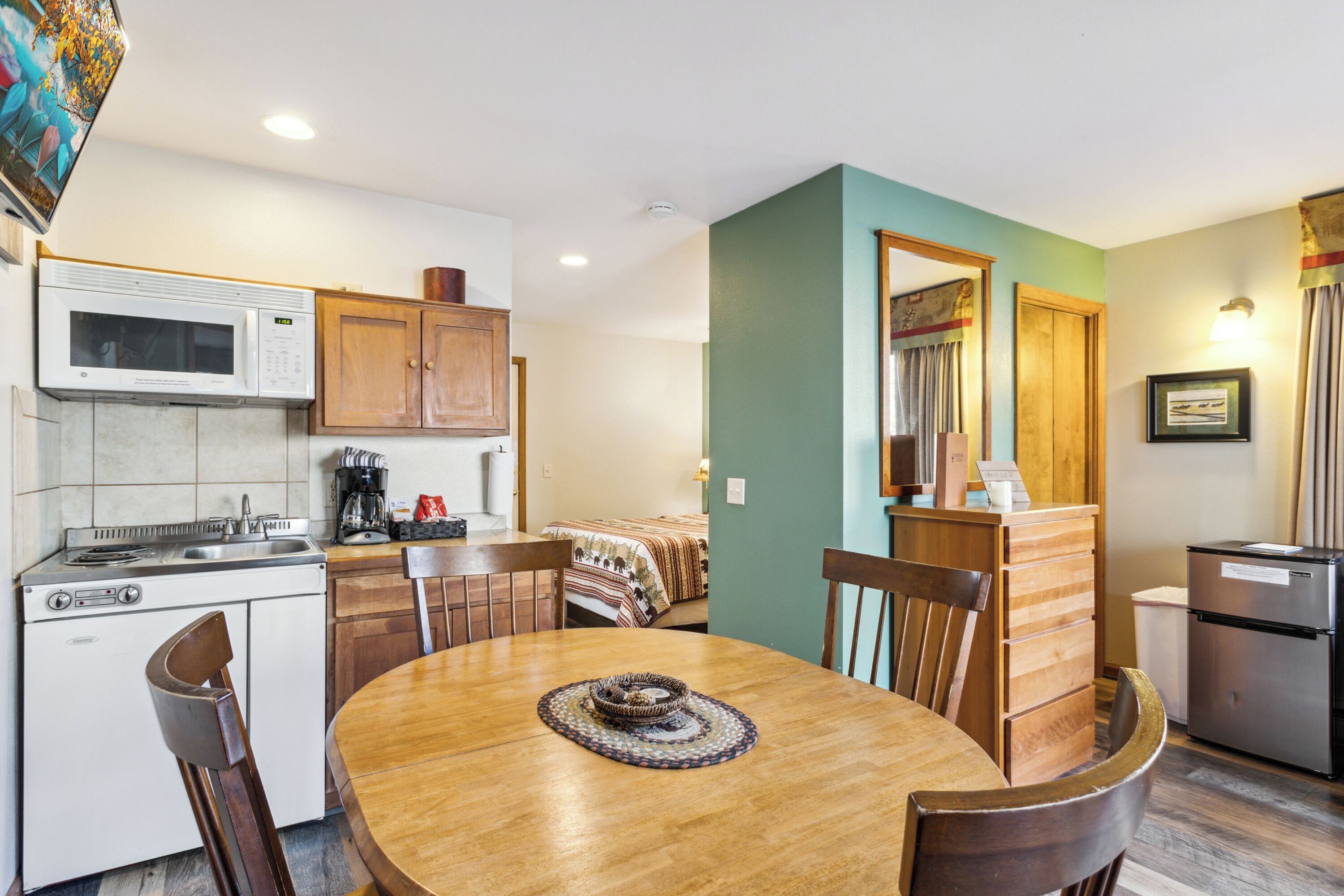 A compact kitchen with a microwave, stove, and sink. There is a dining table with chairs and a small rug. The room has a green accent wall and wooden cabinets.