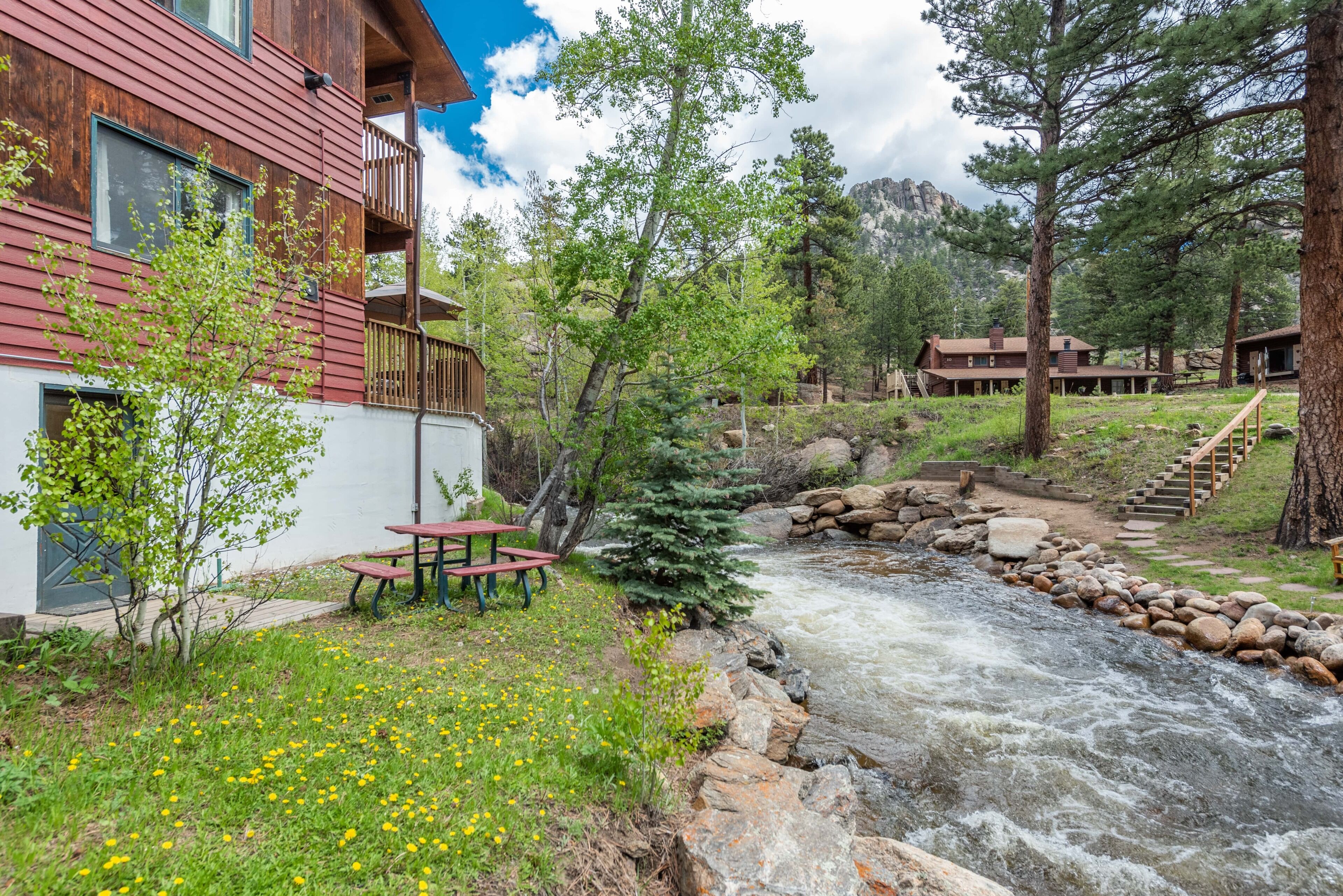 Un chalet rustique avec une façade en bois, un sentier en pierre et une petite table de pique-nique près d’un ruisseau.