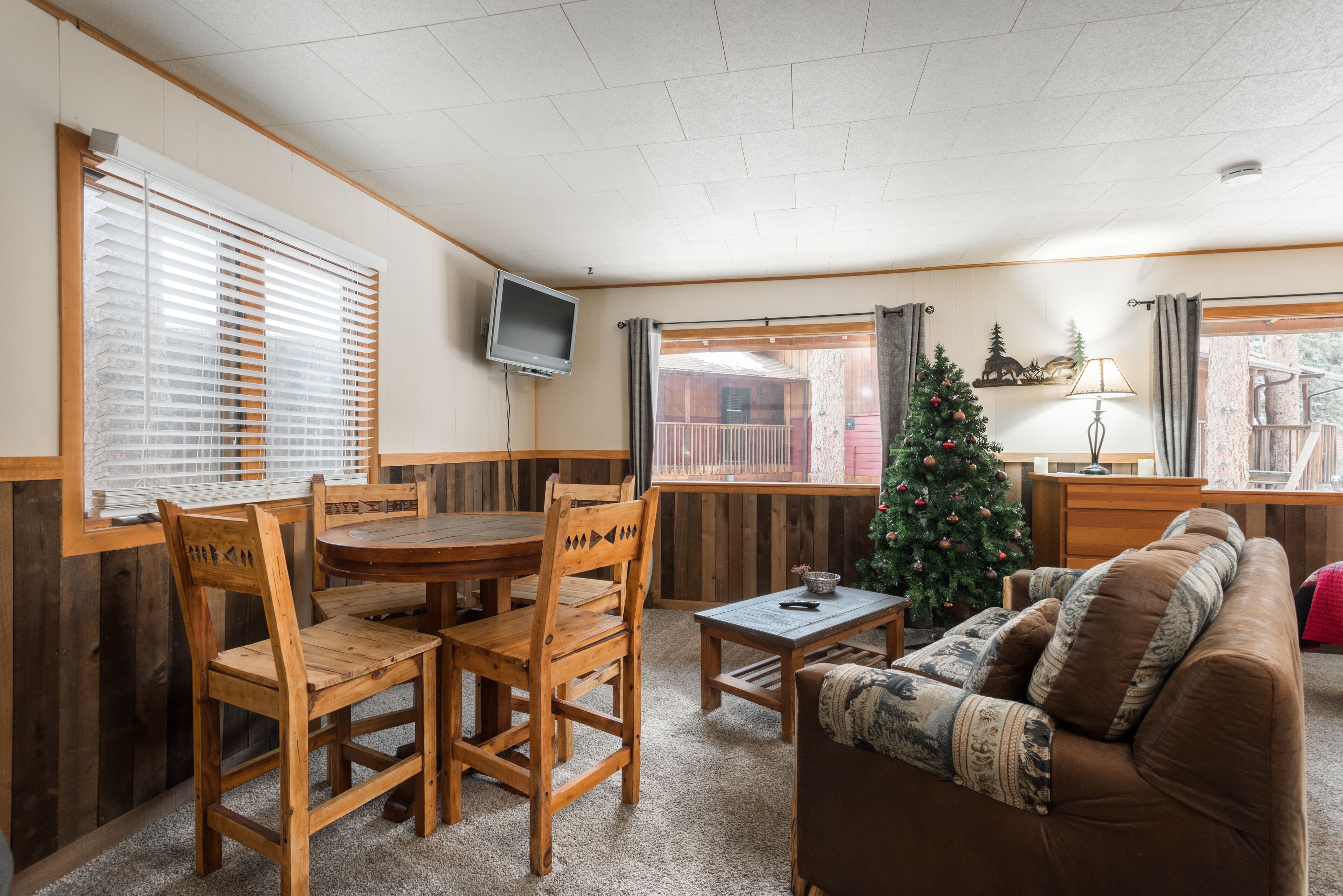 Ein gemütliches Wohnzimmer mit einem Holzesstisch, Stühlen, einem braunen Sofa, einem Weihnachtsbaum und einem Fernseher an der Wand.