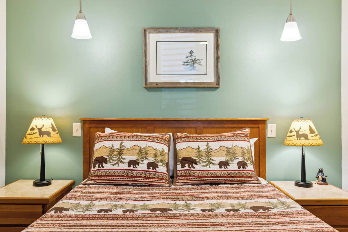 Un letto con copriletto decorato, due abat-jour, un quadro incorniciato al muro e una testiera in legno.