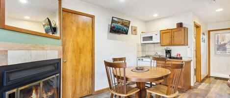 A dining area with a fireplace, wooden chairs, a round table, and a kitchen with a microwave and cabinets.