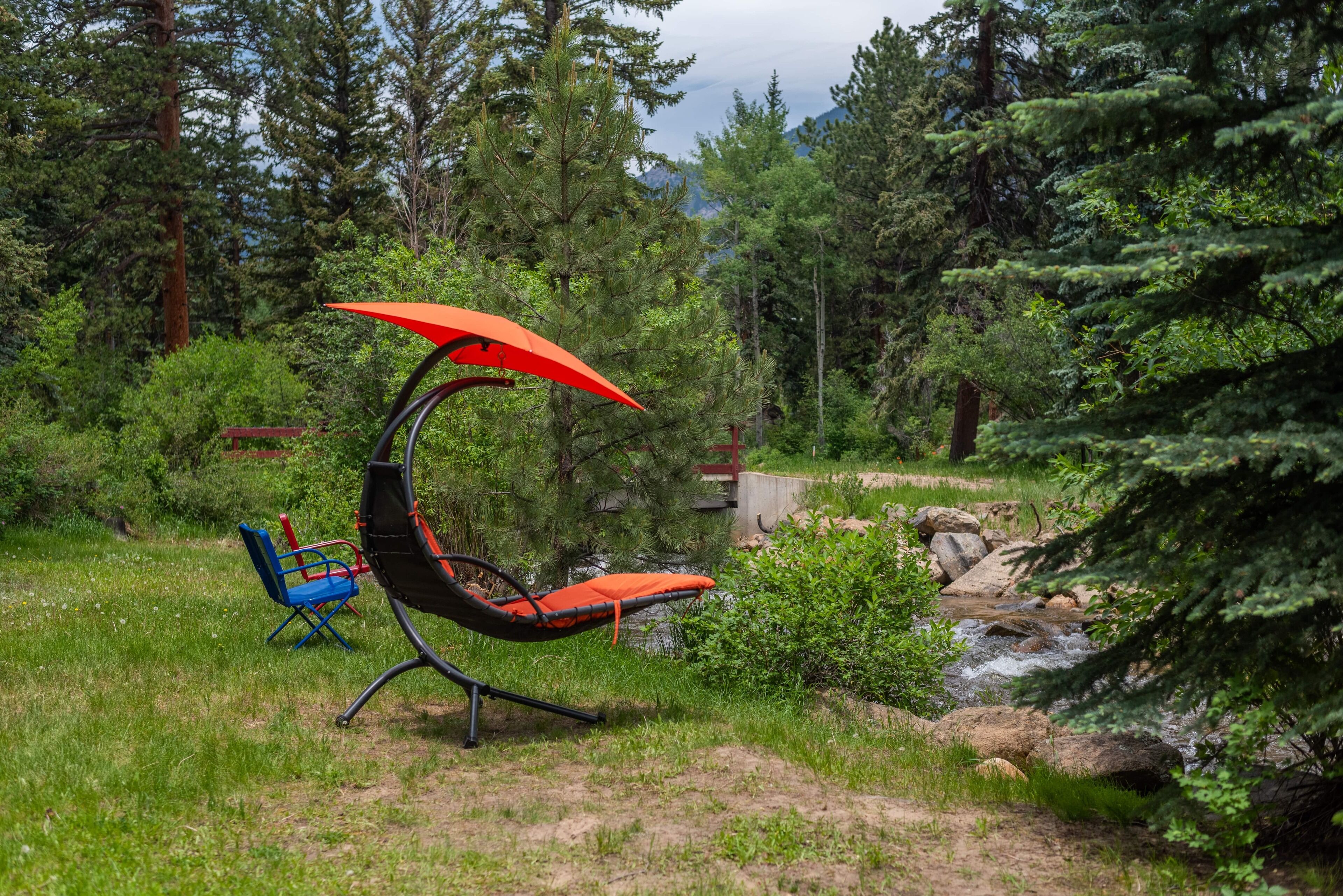 Un fauteuil suspendu en forme de hamac, doté d’une toile orange, installé dans une zone boisée près d’un ruisseau.