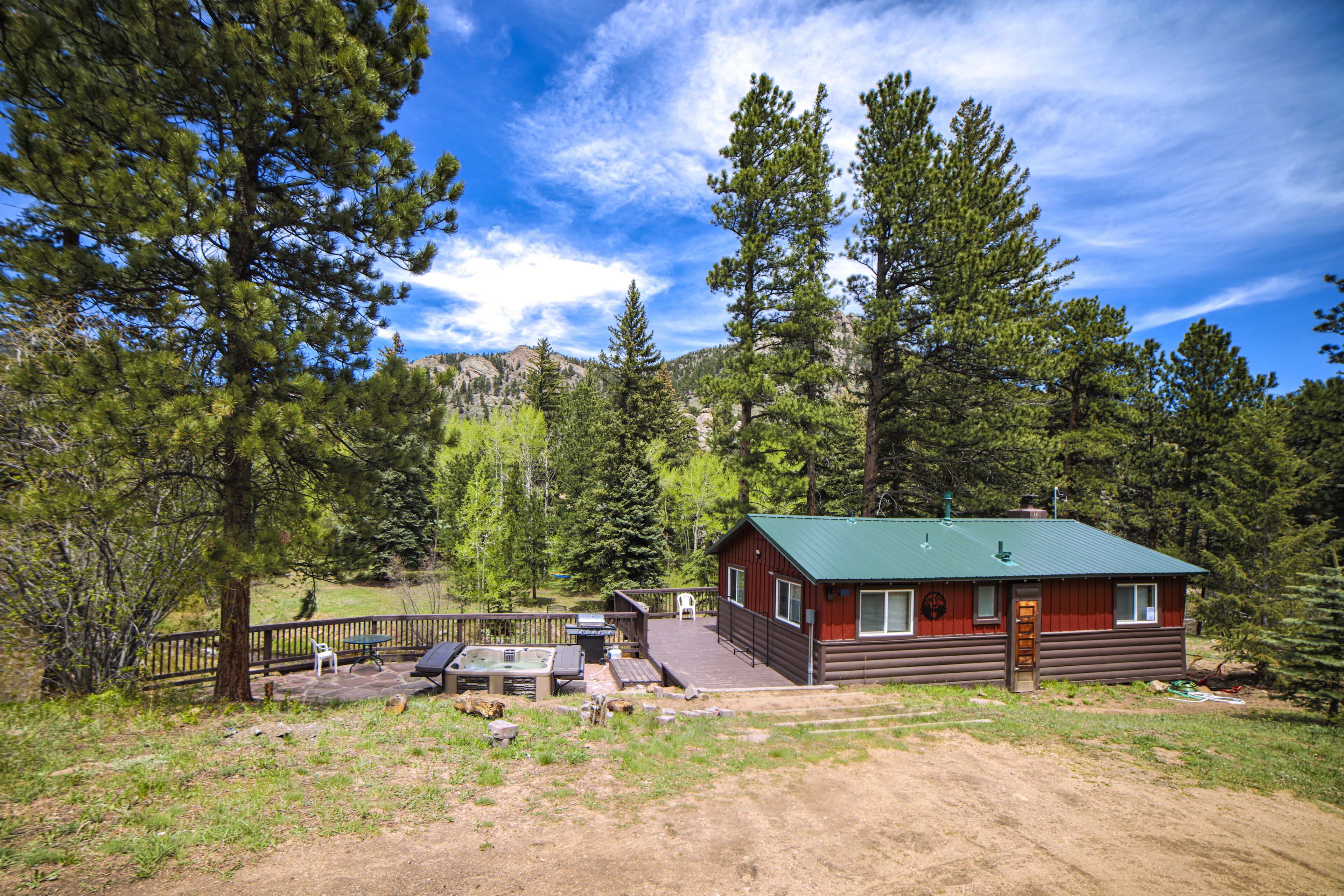 A cabin with a green roof, surrounded by trees, and equipped with outdoor seating and a hot tub.