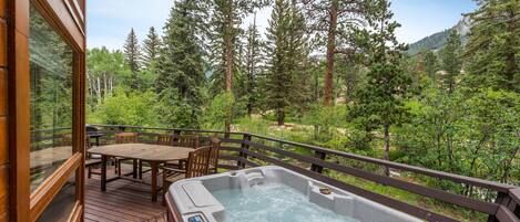 Een houten terras met een jacuzzi, houten meubels en uitzicht op een bebost gebied.