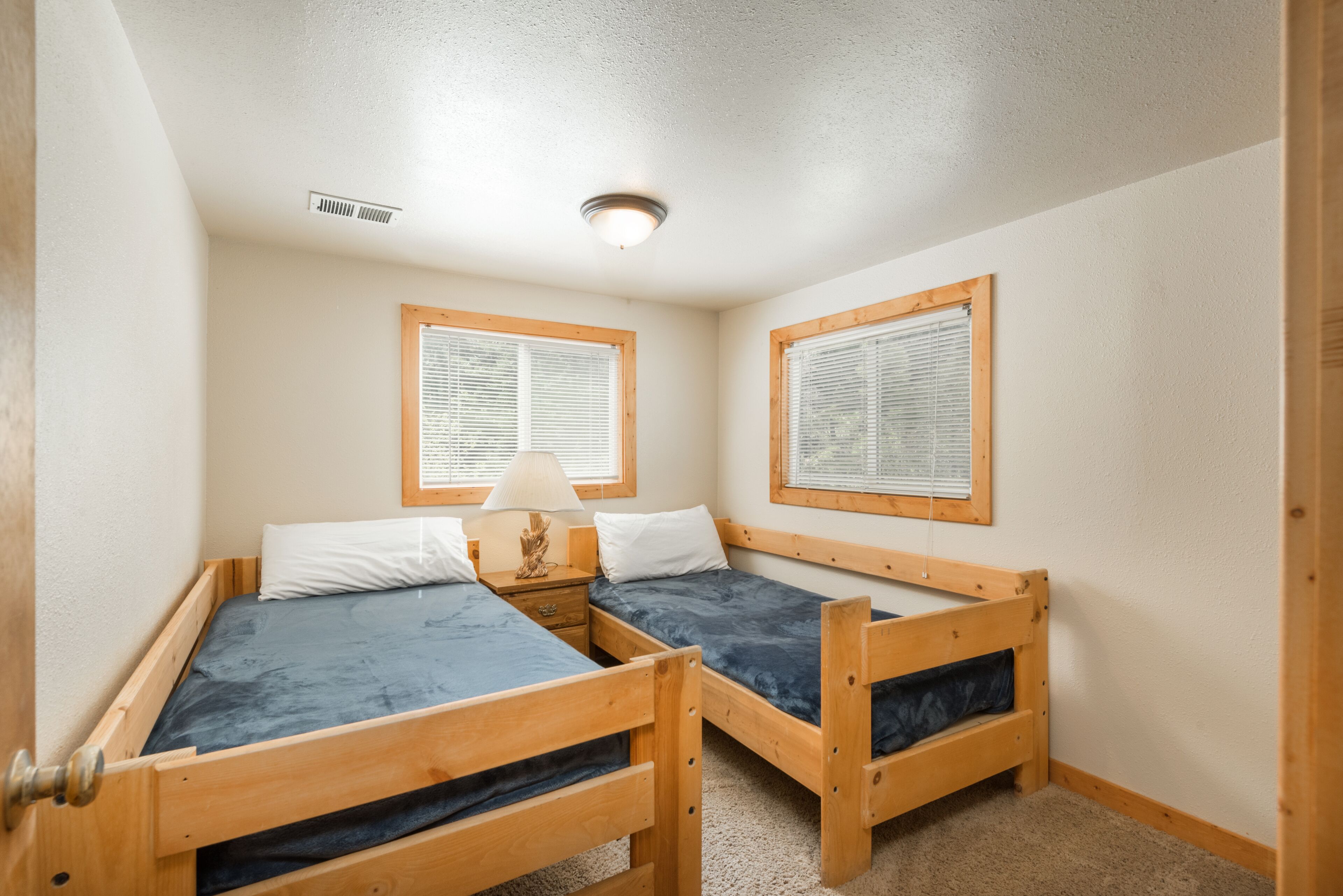 Um quarto pequeno e acolhedor com duas beliches de madeira, uma mesa de cabeceira com abajur e duas janelas com persianas.