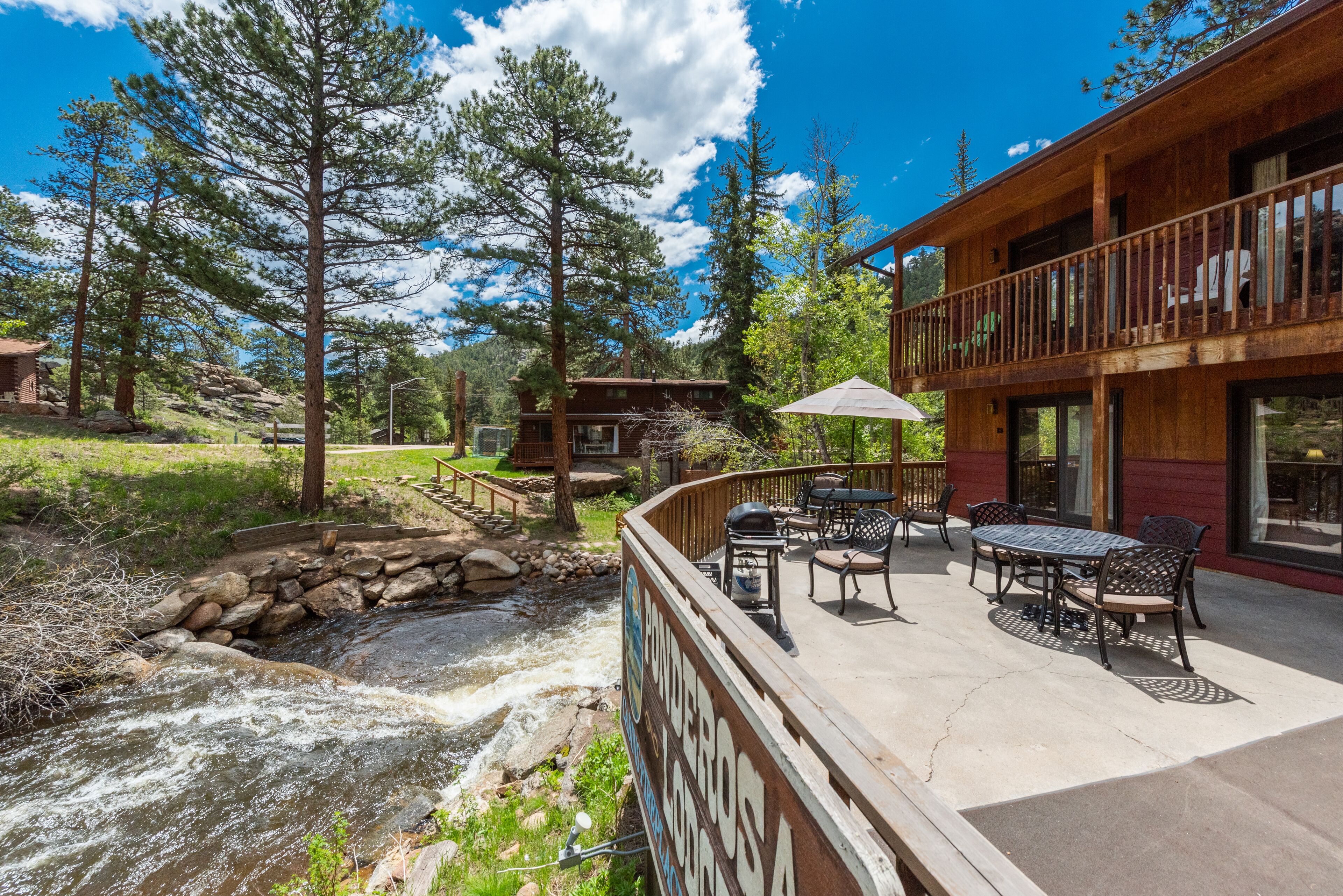 A wooden cabin with a patio overlooking a flowing stream, surrounded by tall trees.