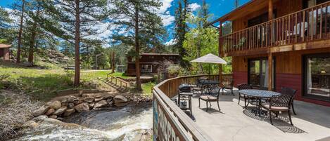 A wooden cabin with a patio overlooking a flowing stream, surrounded by tall trees.
