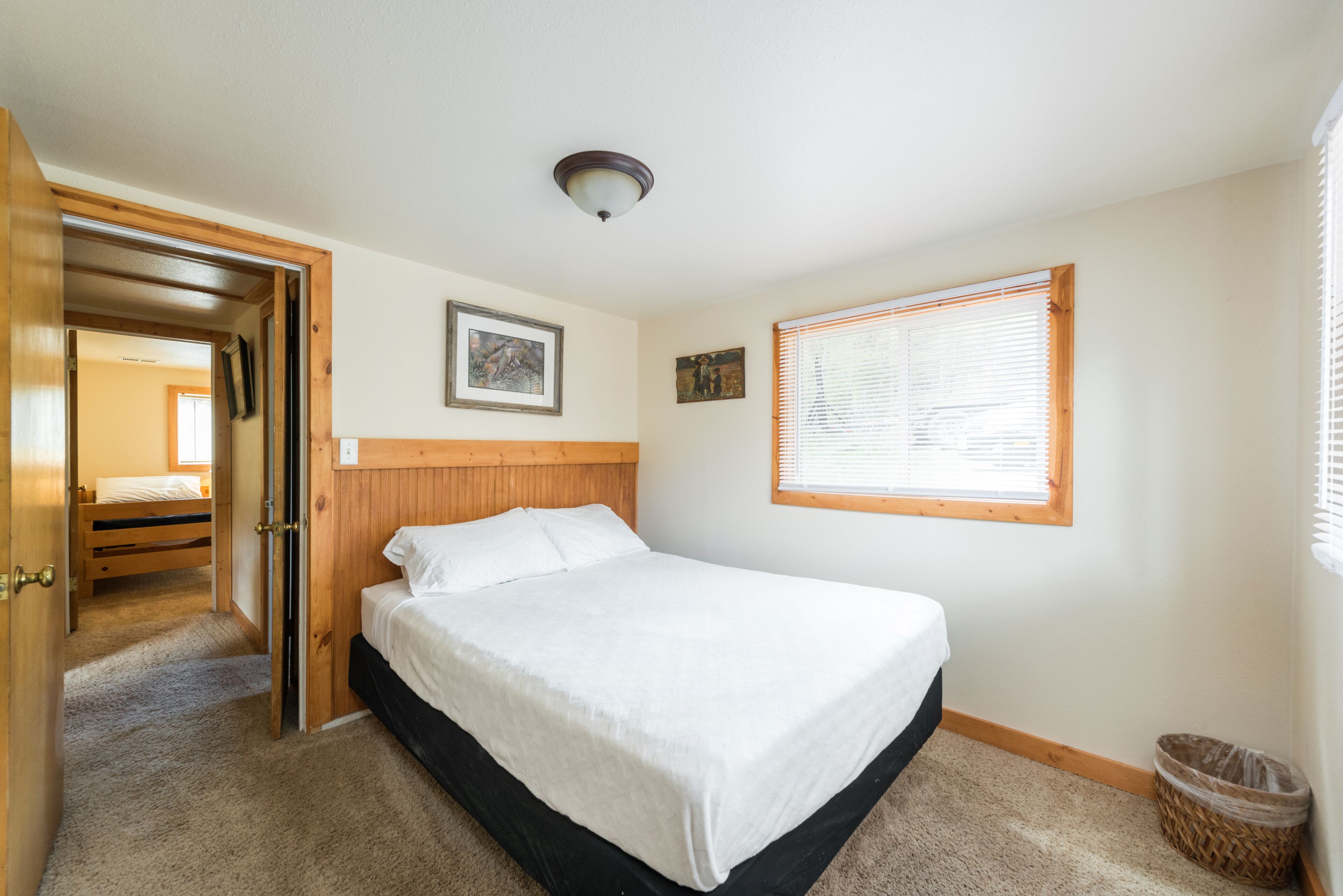 Une chambre à coucher avec une tête de lit en bois, un lit simple, une fenêtre avec des stores et une porte donnant sur une autre pièce.