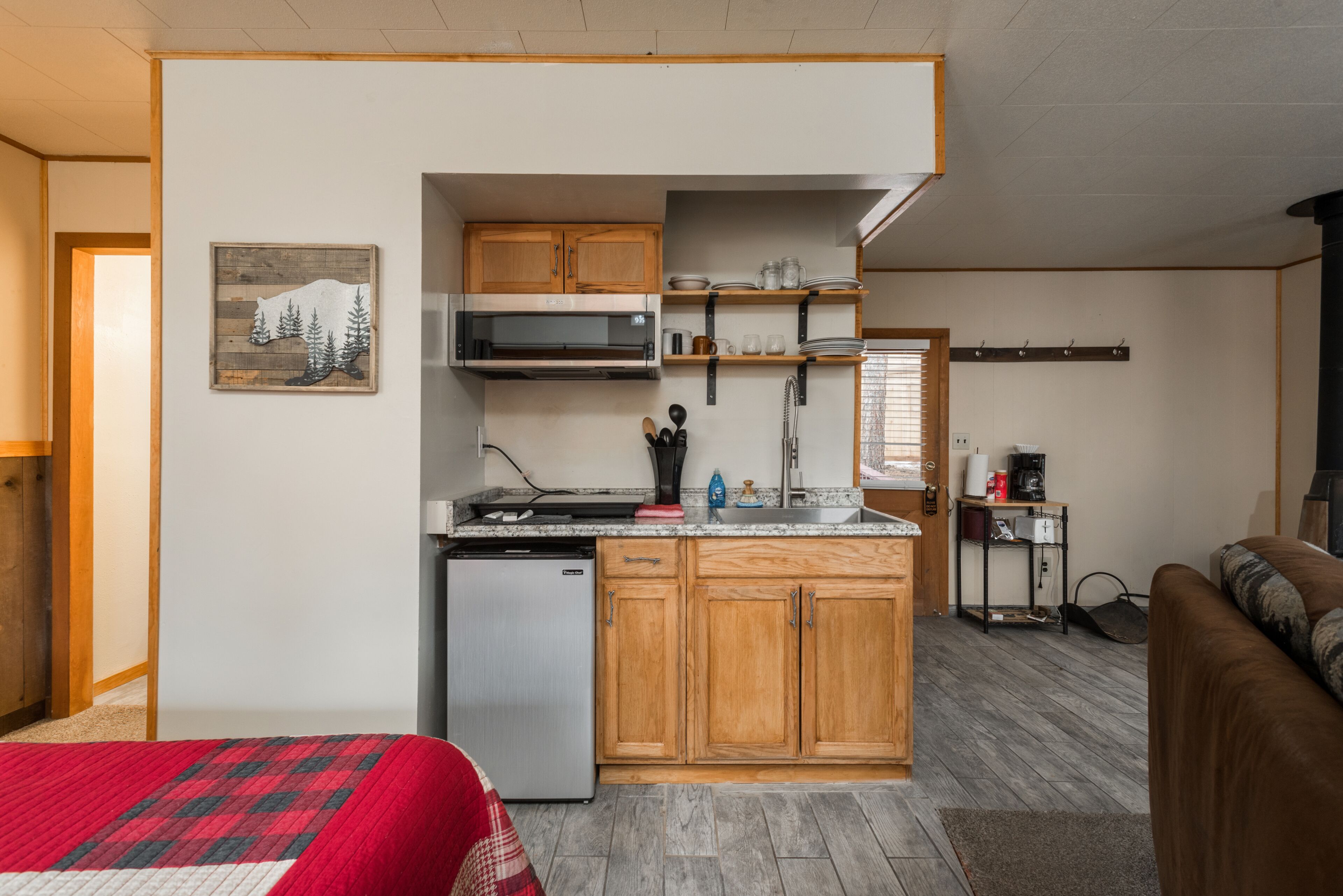 A compact kitchen with wooden cabinets, a microwave, and a dishwasher. There is a bed with a red and white blanket, and a brown couch in the living area.