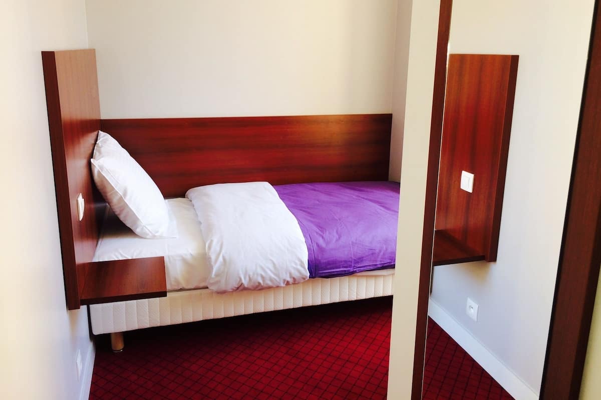 A single bed with a wooden headboard, white and purple bedding, and a red patterned floor.