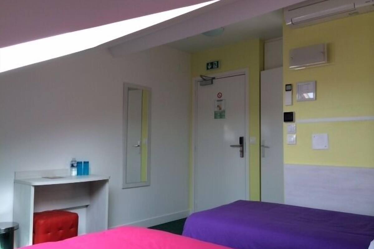 A room with two beds, a desk, and a chair. There is a door with a sign above it and a light switch on the wall.
