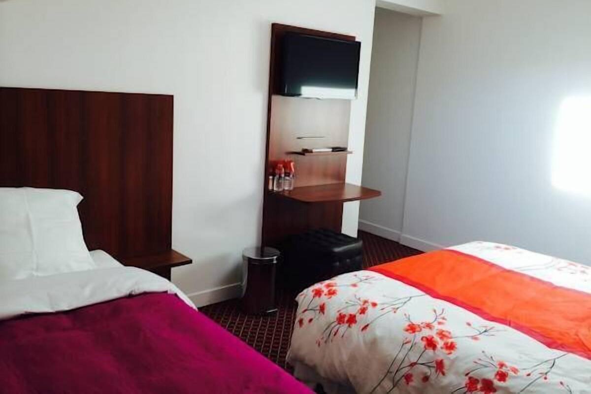 A hotel room with a wooden headboard, a bed with a red blanket, a white bedspread with red flowers, a small wooden shelf with toiletries, and a flat-screen TV mounted on the wall.