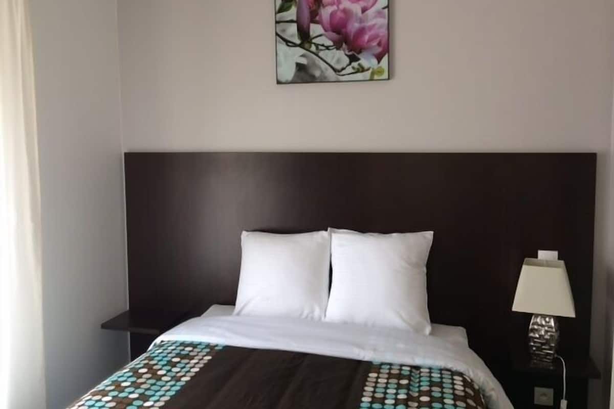 A hotel room with a dark wood headboard, white bedding, a patterned throw blanket, a painting of pink flowers on the wall, and a bedside lamp.