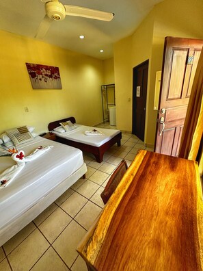 A hotel room with two beds, a wooden dining table, a ceiling fan, a painting on the wall, and a door with a number sign. - Hotel Nany (Cabo Velas)