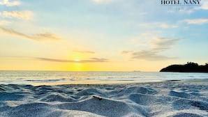 A beach with pebbles and waves, a sunset in the background, and a logo for HOTEL NANY in the top right corner. - Hotel Nany (Cabo Velas)