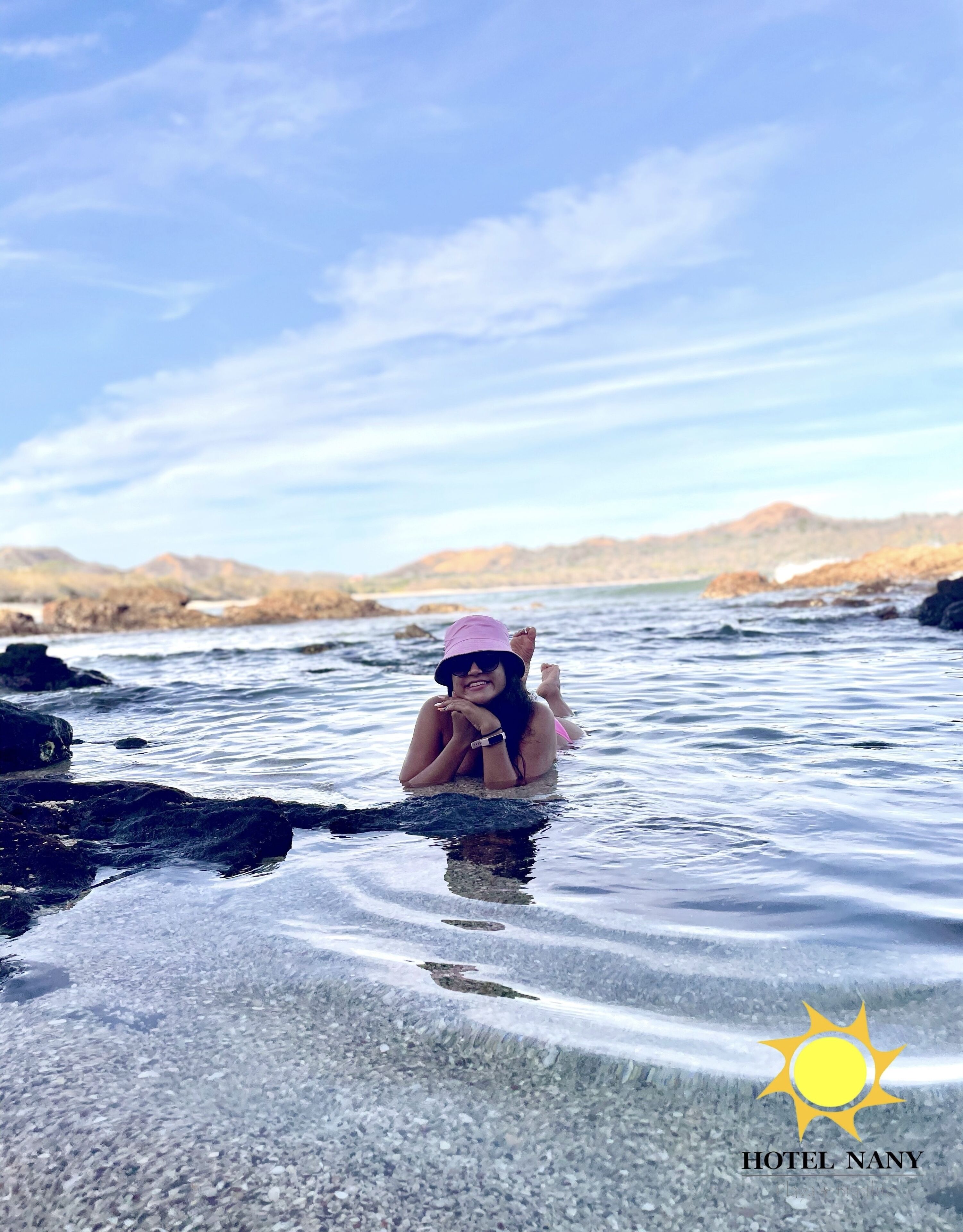 A person in a clear pool with a rocky shoreline and distant hills.