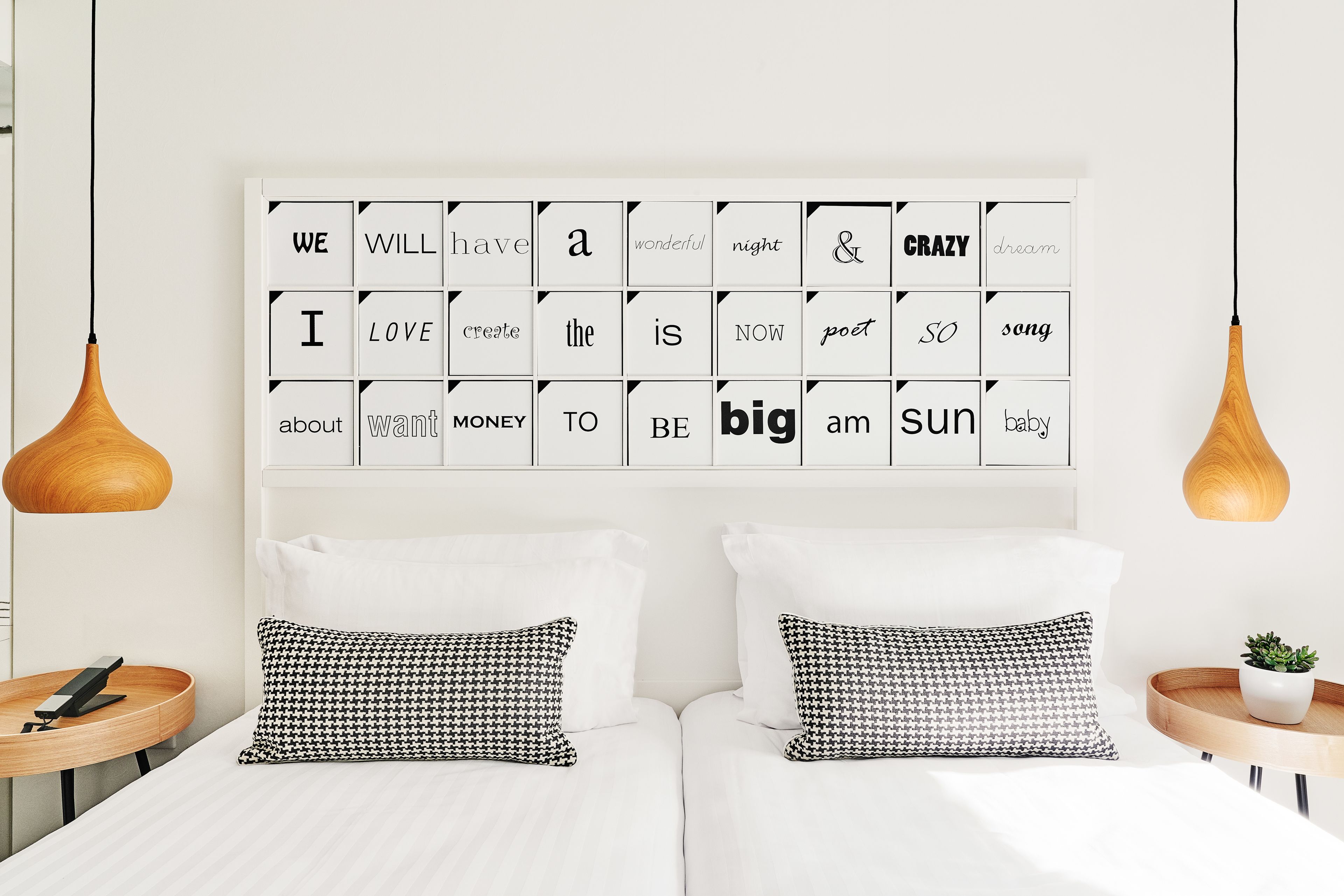 Quarto moderno com cama branca, almofadas xadrez, uma obra de arte na parede com frases inspiradoras e luminárias pendentes.