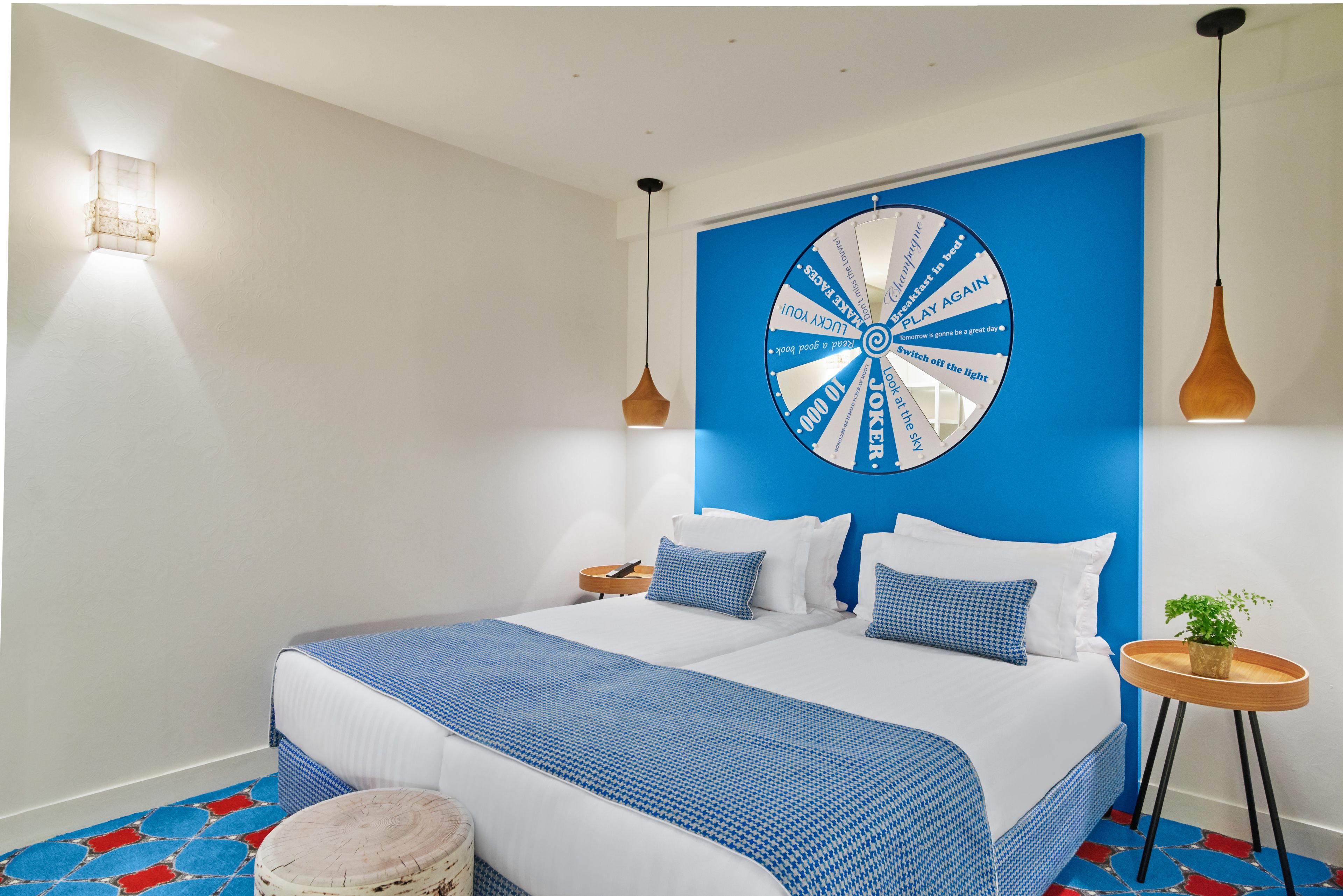 A modern hotel room with a large bed, a blue accent wall, a wall-mounted circular light fixture, and a small round table with a plant.