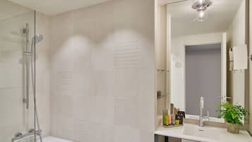 Un baño moderno con ducha envuelta en vidrio, bañera blanca, espejo grande y lavamanos con artículos de aseo.