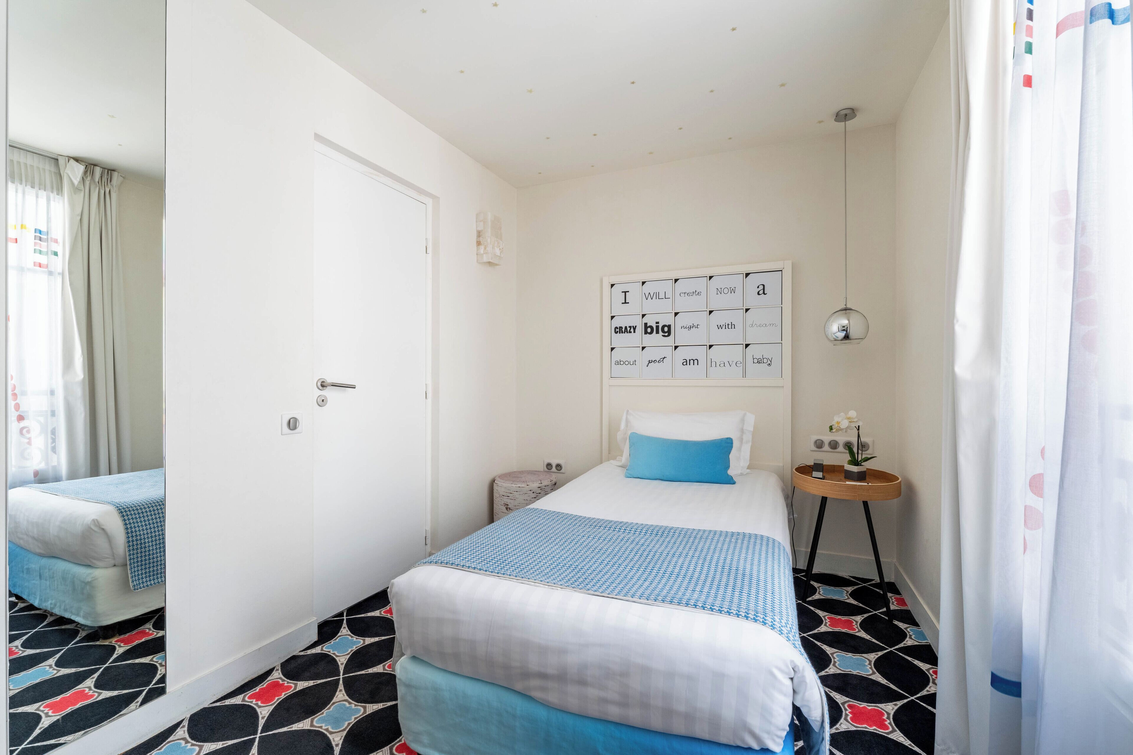 A hotel room with a bed, a bedside table with a lamp, a wall with motivational quotes, and a patterned floor.