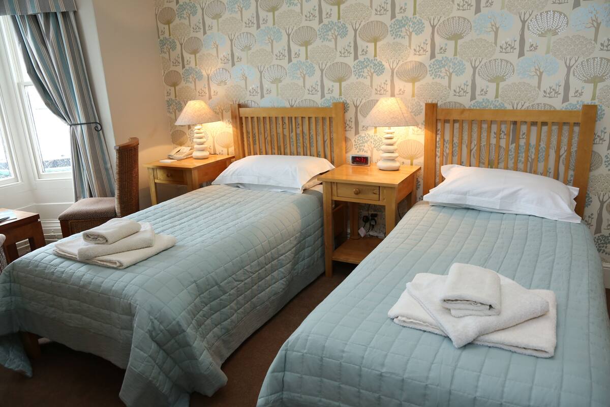 A hotel room with two beds, each with a wooden headboard and a bedside table with a lamp. There is a window with curtains, a chair, and a small table with a phone and a lamp.