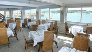 A restaurant with tables set for dining, wicker chairs, and large windows offering a view of boats on the water.