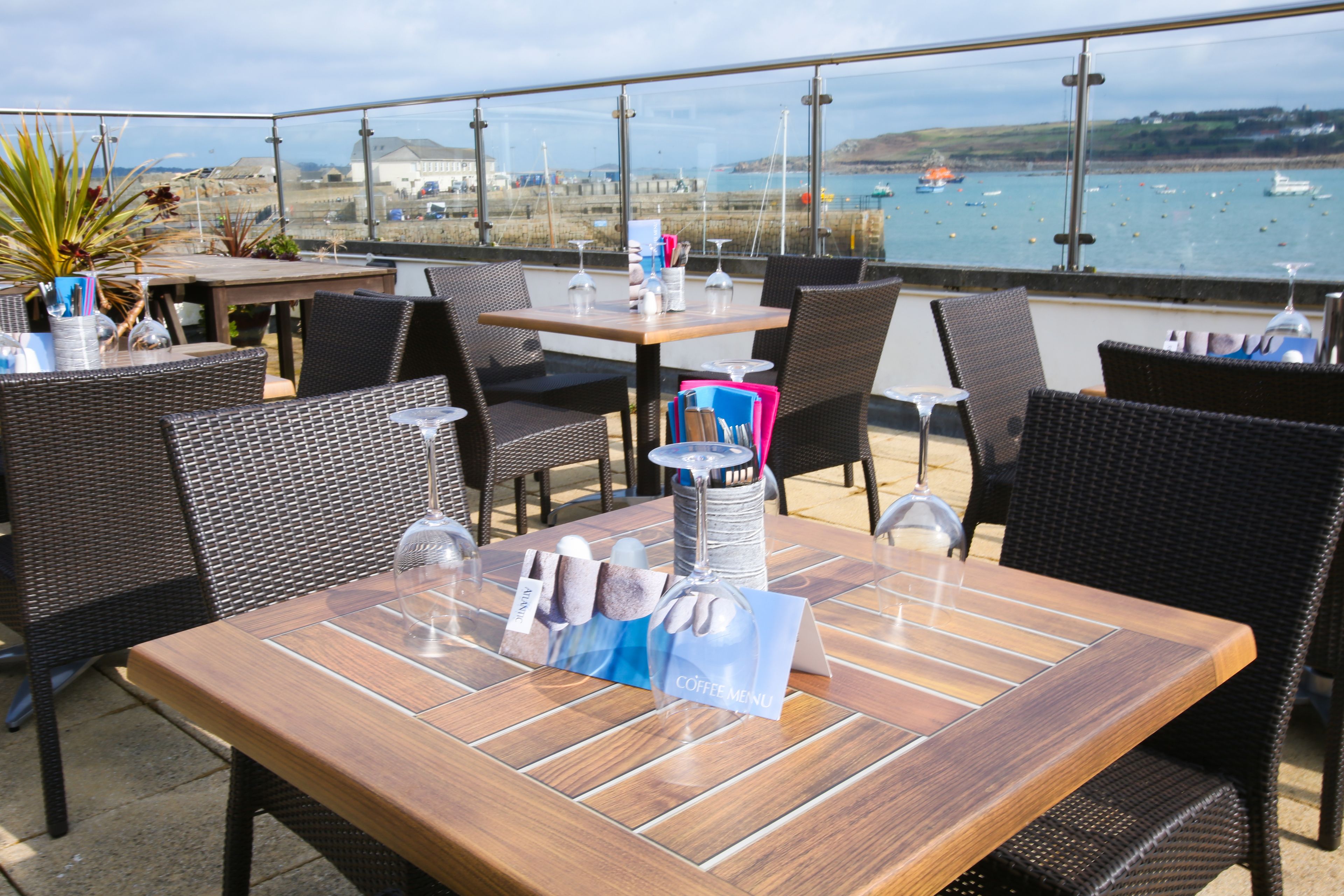 An outdoor dining area with wicker furniture and a wooden table, overlooking a coastal view with boats and buildings.