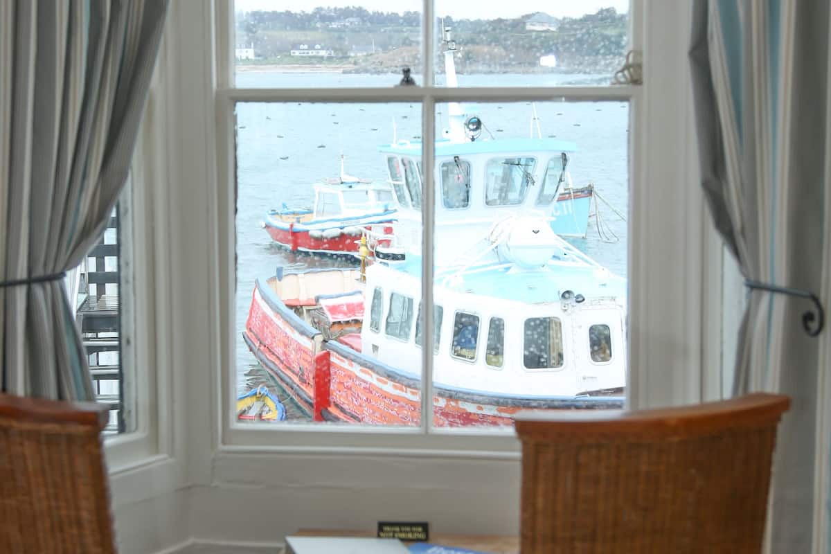 A room with a large window overlooking a harbor, featuring boats and a dock. There are two wicker chairs and a small table with a book.