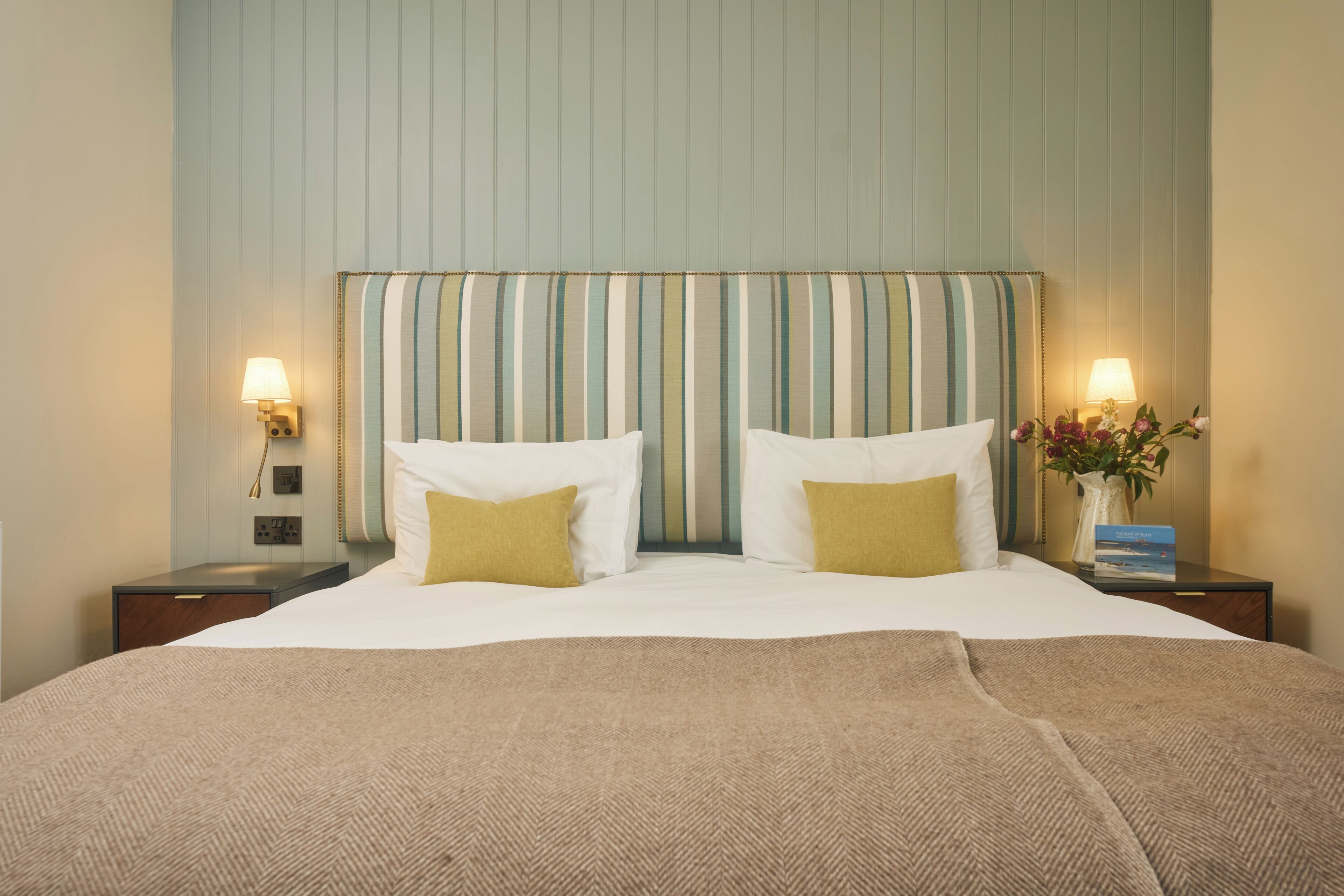 A hotel room with a large bed, two bedside lamps, a nightstand with a vase of flowers, and a striped headboard.