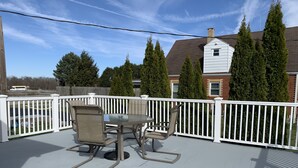 Une terrasse couverte avec une table et des chaises, des rampes blanches et une vue sur un quartier résidentiel avec des arbres et des maisons.