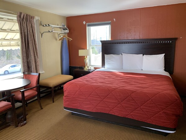 A hotel room with a large bed, a small table, a chair, a bench, a window with curtains, and a view of the outside.