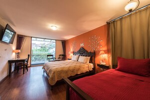 Habitación de hotel con una cama grande, otra más pequeña, un escritorio con silla, un televisor montado en la pared y un balcón con vistas a edificios.