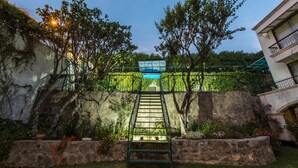 Una escalera de piedra conduce a un puente de cristal, rodeado de abundante vegetación y plantas con flores.