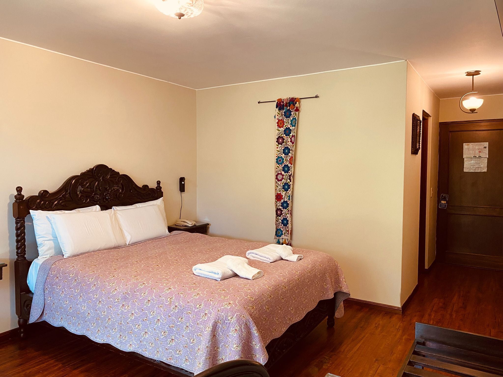 A bedroom with a wooden bed, a bedside table with a lamp, a hanging floral curtain, and a door with a lock.