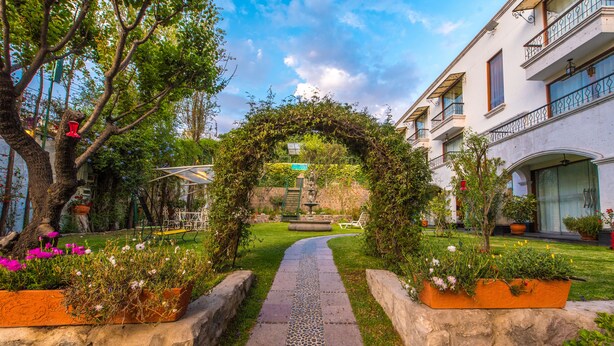 Un camino serpentea por un jardín con borde de piedra, rodeado de follaje y plantas en macetas.