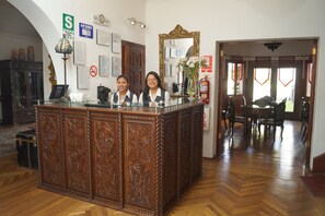 Un mostrador de recepción con paneles de madera decorativos, un espejo y un jarrón con flores. Hay un cartel con la letra 'S' y un letrero de prohibido fumar en la pared.