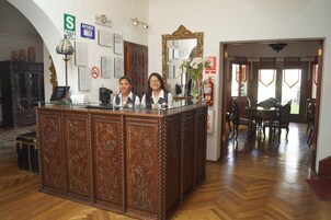 Un mostrador de recepción con paneles de madera decorativos, un espejo y un jarrón con flores. Hay un cartel con la letra 'S' y un letrero de prohibido fumar en la pared.