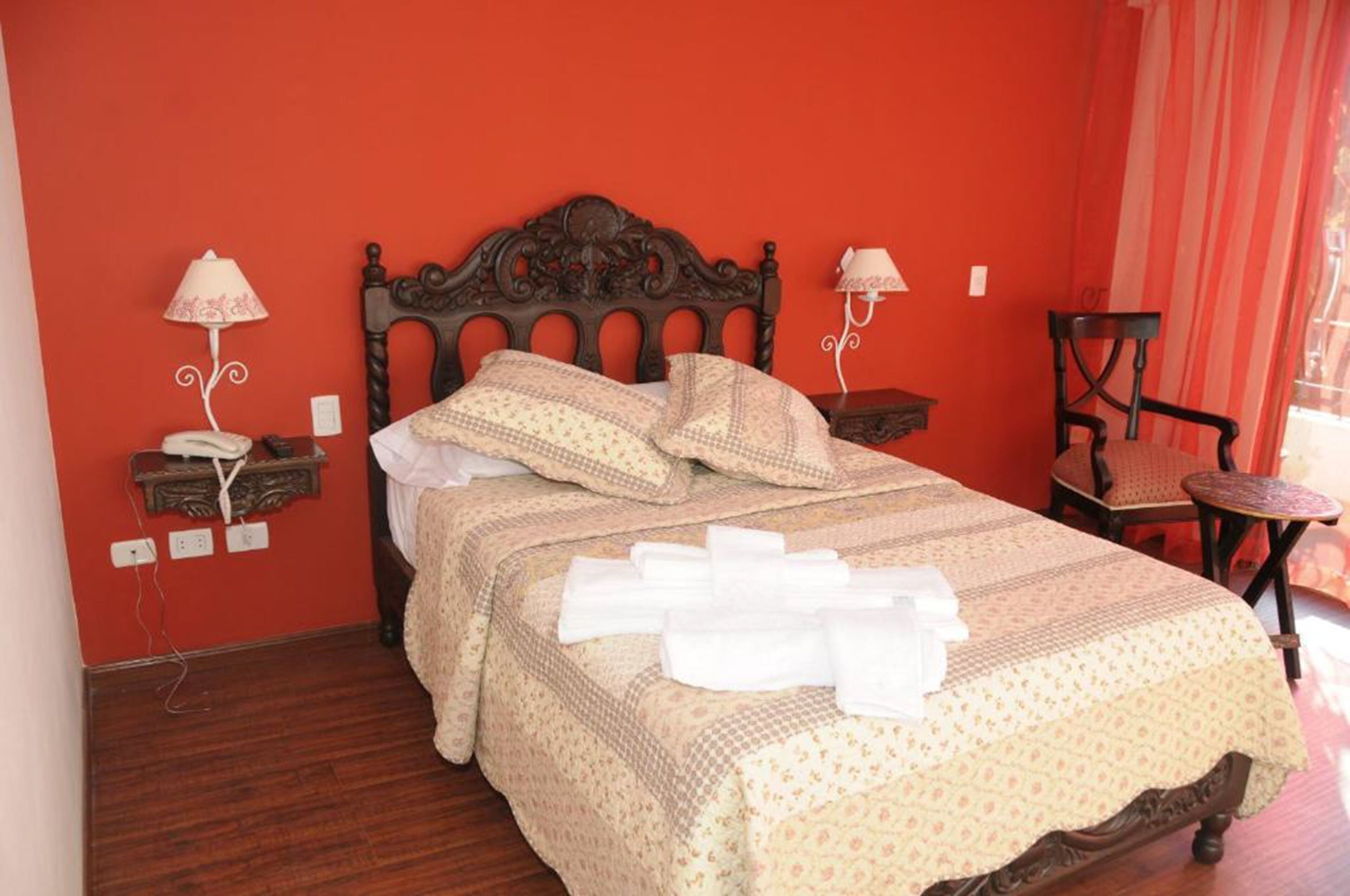 A neatly made bed with a patterned bedspread and pillows, two bedside lamps, a chair, and a small table in a room with red walls and wooden flooring.