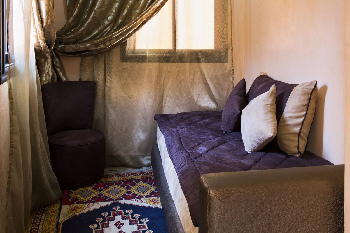 Um quarto pequeno e aconchegante com uma cama, uma cadeira e uma janela com cortinas.