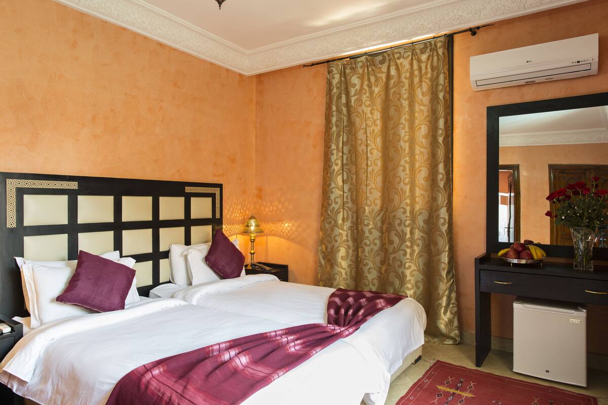 Quarto de hotel com duas camas, uma escrivaninha com espelho, ar condicionado e um pequeno frigorífico.