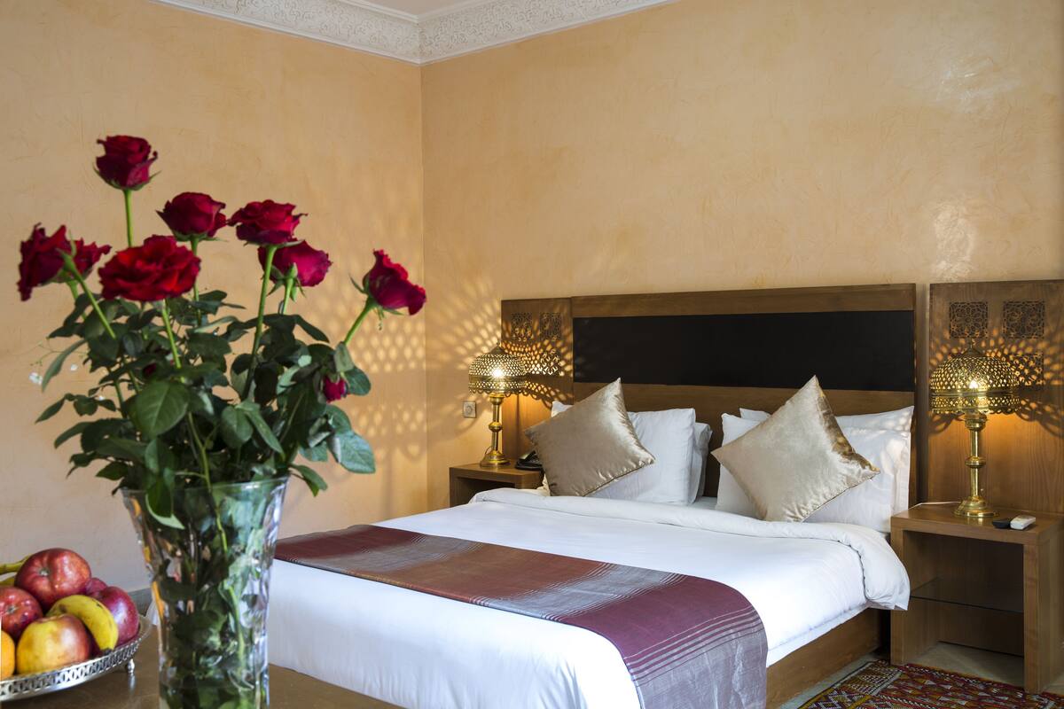 Quarto de hotel com uma cama, abajures, um vaso com rosas e uma fruteira.
