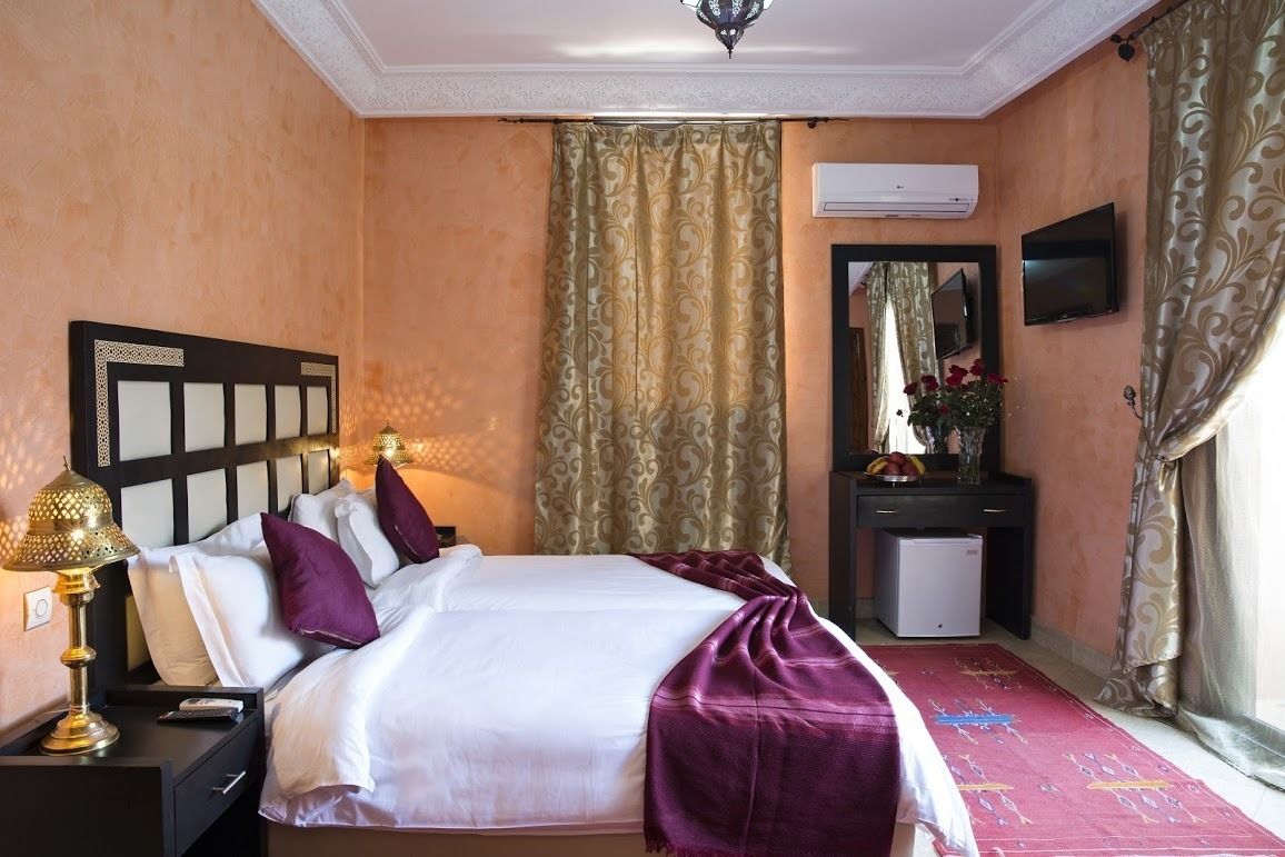 A hotel room with a bed, nightstand, lamp, air conditioning unit, TV, and a small desk with a mirror.