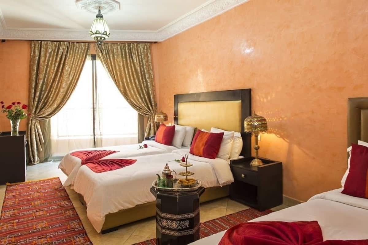 Quarto de hotel com duas camas, um candelabro, uma mesa de cabeceira com abajur, um vaso com flores e vista para o exterior.