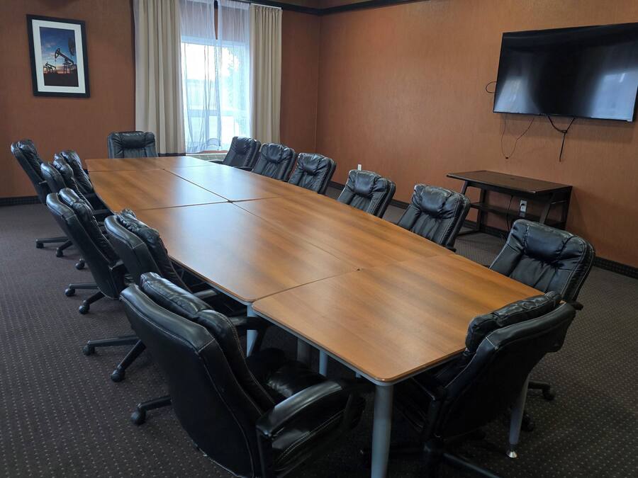 A conference room with a long wooden table surrounded by black leather chairs, a television on the wall, and a window with curtains.