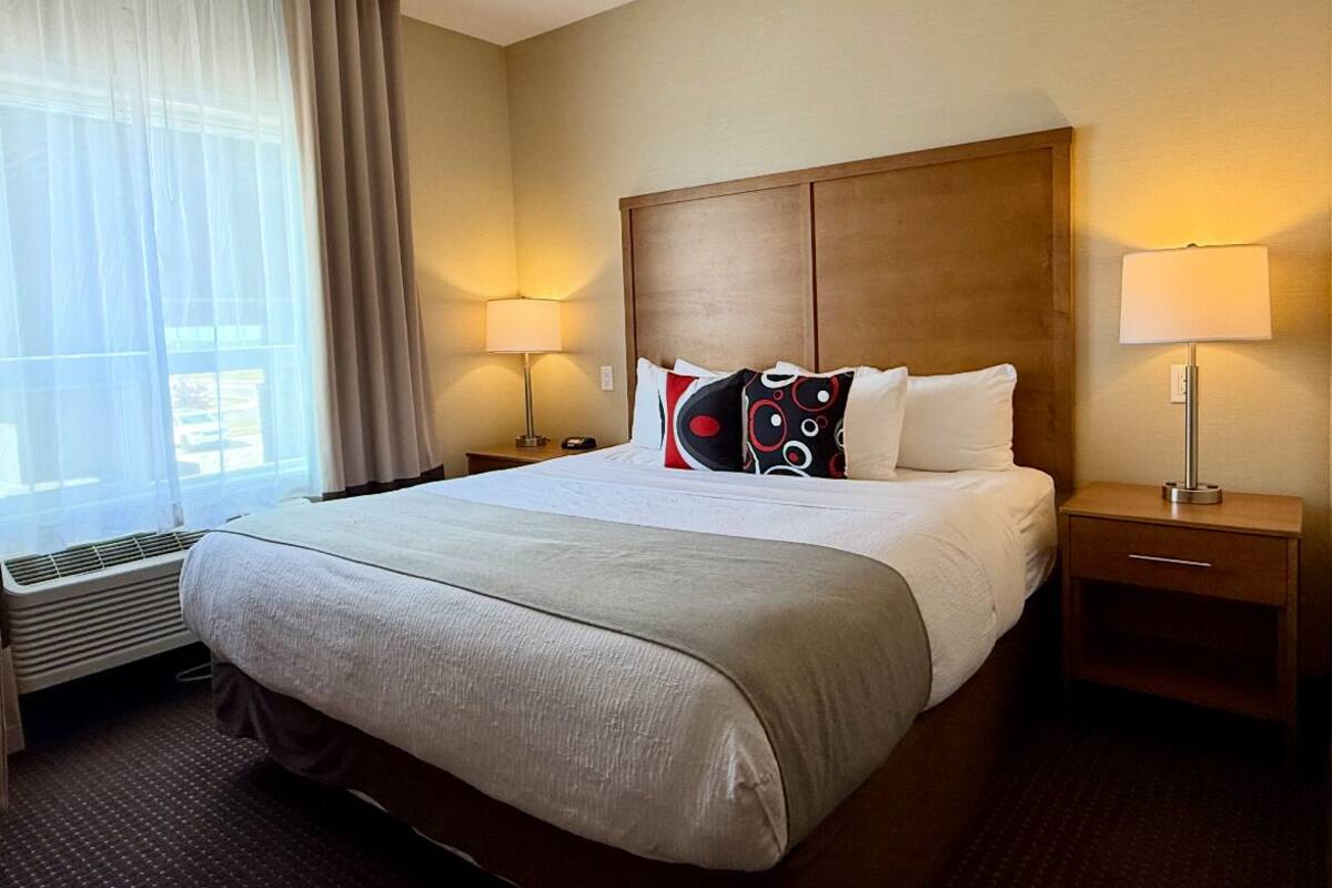 Une chambre d’hôtel avec un grand lit, une tête de lit en bois, deux tables de chevet avec des lampes, une fenêtre avec des rideaux légers et un climatiseur fixé au mur.