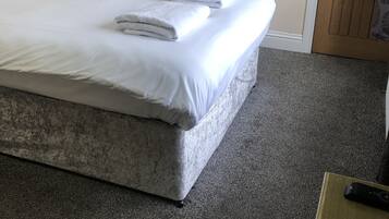 Uma cama bem arrumada com roupa de cama e toalhas brancas, uma pequena mesa de cabeceira com abajur, uma luminária na parede e uma porta com espelho decorativo.
