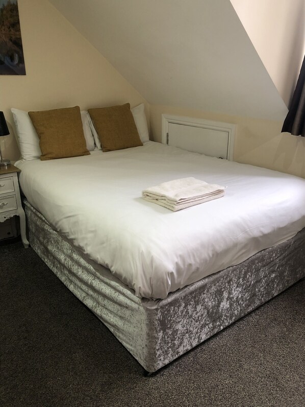 A neatly made bed with white linens and two brown pillows, a white bedside table with a lamp, and a dark carpeted floor.