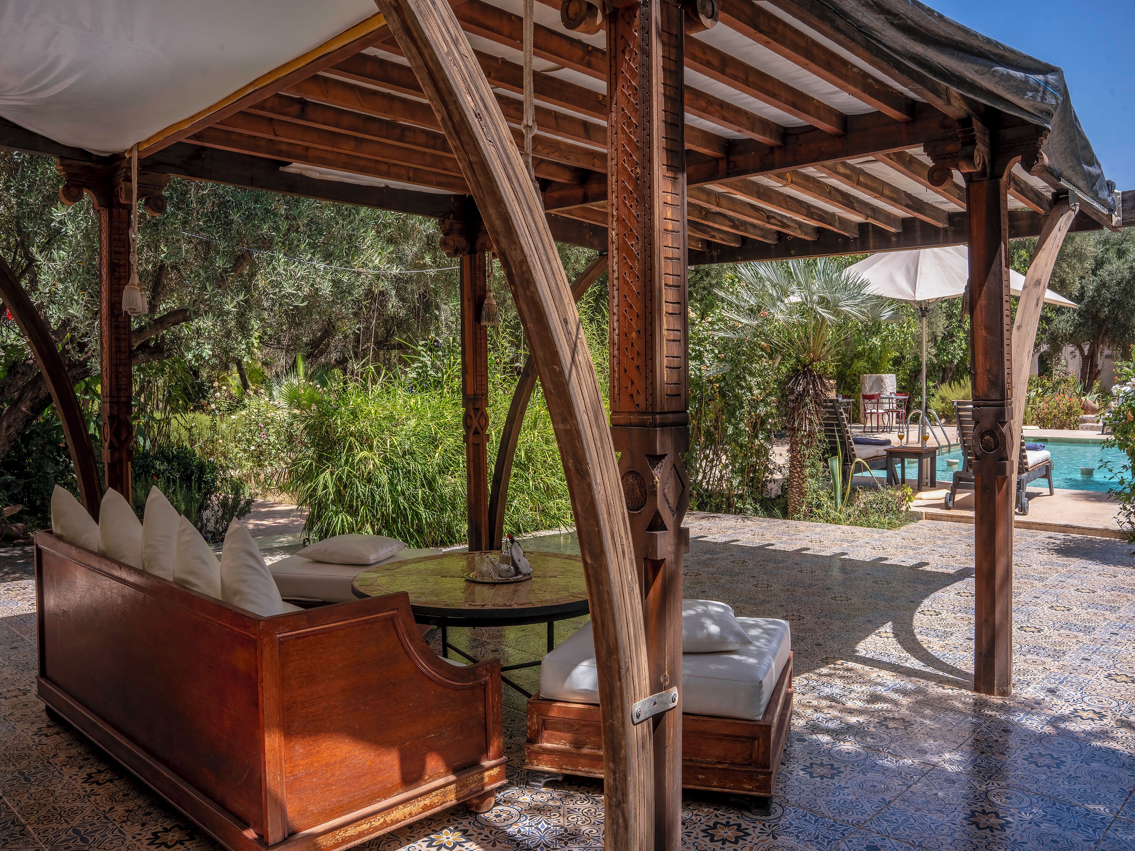Un espacio al aire libre cubierto con un banco de madera y un sofá acolchado, rodeado de abundante vegetación y una piscina.