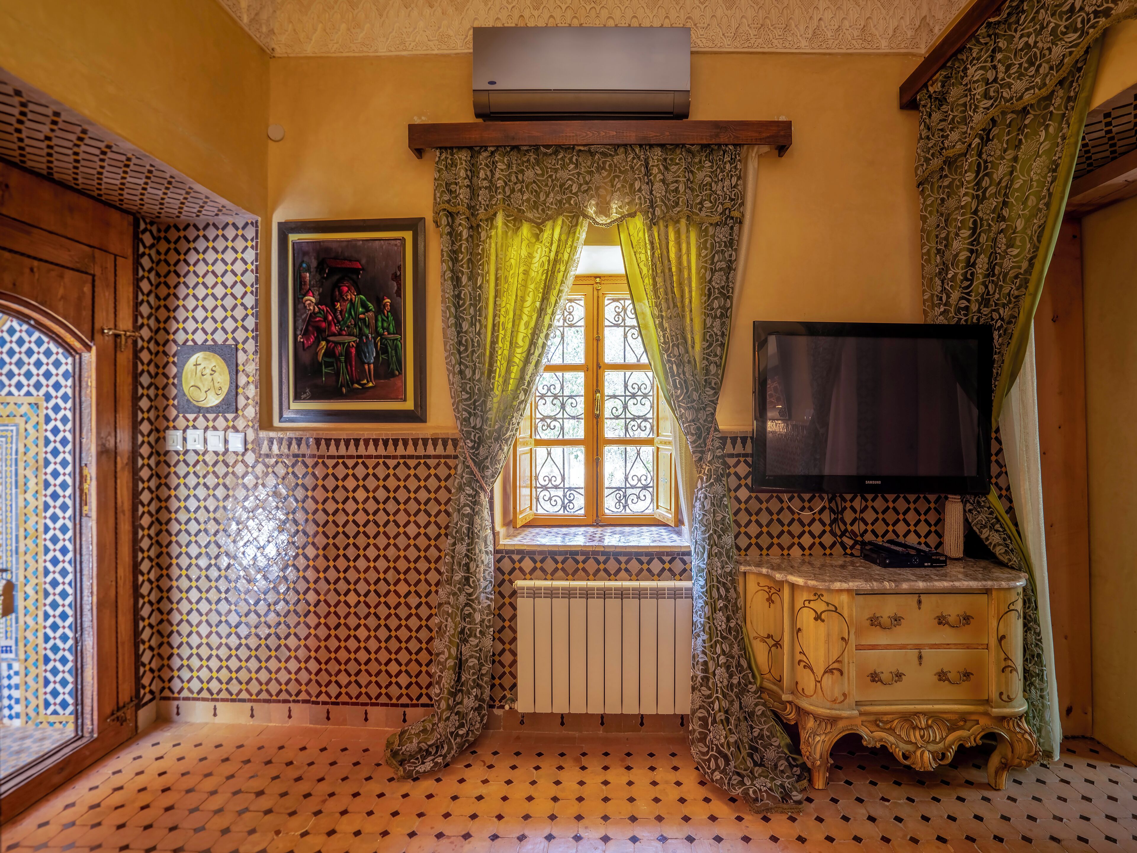 Ein Zimmer mit Wandfliesen mit Muster, einem Fenster mit dekorativen Vorhängen, einem Heizkörper, einem Fernseher und einem Holzschrank.